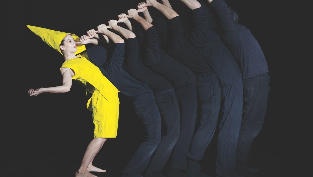 A women in a yellow outfit with a thumb to her nose with other dancers in black outfits doing the same in front of her