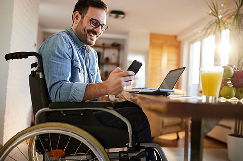 Image entitled man in wheelchair sitting at the table with his laptop on the table and mobile phone in his hands